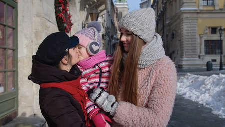 Lovely sisters holding younger sister child girl in hands. Tourists family talking, smiling, embracing in old city. Travelers enjoy time together on Christmas winter holiday vacation on town streetの写真素材