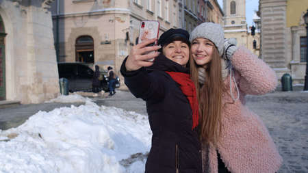 Two young smiling women tourists bloggers taking selfie photos portrait, video conferencing call on mobile phone on city street. Happy lesbian LGBT couple embracing. Winter holidays traveling vacationの写真素材
