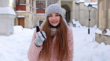 Cheerful young woman looking to camera, showing thumb up gesture sign while standing outdoor in...