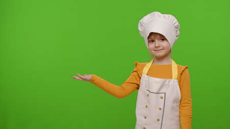 Cute child girl dressed in apron and hat like chef cook pointing at left on blank space. Place for your advertisement . Chroma key background. Nutrition, family cooking school, children educationの写真素材