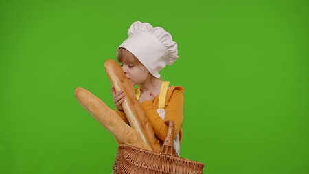 Little child girl dressed in apron and hat like chef cook showing basket with baguette and bread,...