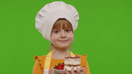 Portrait of little child girl kid dressed as professional cook chef baker eating tasty strawberry cake isolated on chroma key background. Concept of sweet nutrition food, cooking school, educationの写真素材