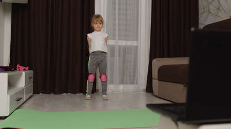 Little children girl watching online distance group lessons with trainer on laptop, exercising at home. Child kid doing gymnastics stretching pilates workoutの写真素材
