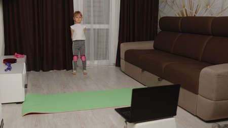 Kid girl doing sport exercises, watching video lesson online on laptop computer at home. Sport...