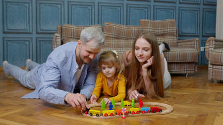 Family of mother, father with daughter child girl riding toy train on wooden railway at home room