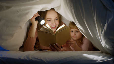 Mother and daughter lying in bed at home under duvet blanket and reading bedtime stories, fairytalesの写真素材