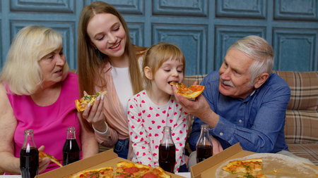 Multigenerational family having lunch party, feeding child girl with pizza, laughing, enjoying meal