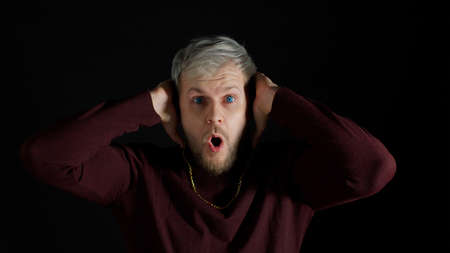 Shocked amazed handsome man in stylish blouse feeling excitement, looking at camera and holding hands near face on black background. Facial expression of trendy bearded guy surprised say wow omgの写真素材