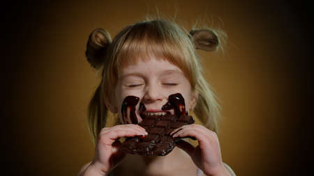 Close-up portrait of teen smiling child kid smears her face with melted chocolate and showing thumbs up sign. Satisfied young blond girl with two ponytails looking at camera on dark brown backgroundの写真素材