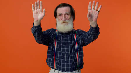 Happy friendly elderly bearded man waves hand palms in hello gesture welcomes someone with...