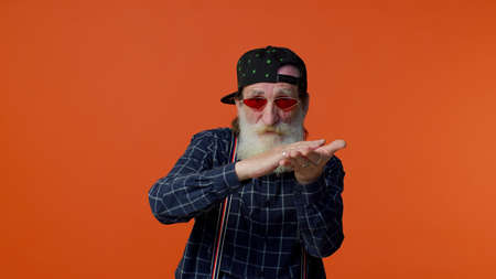Mature old bearded grandfather in sunglasses and cap showing wasting or throwing money around hand gesture, more tips dreaming about big profit body language. Senior man isolated on orange backgroundの写真素材
