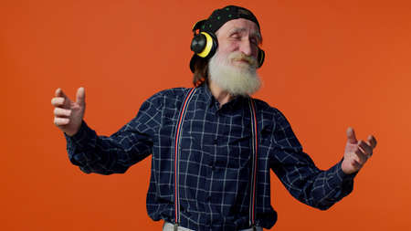 Senior old bearded stylish man listening music via headphones and dancing disco fooling around...