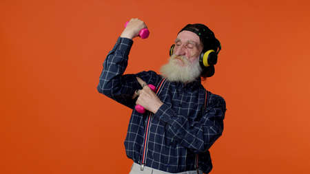 Elderly bearded gray-haired man listening music via headphones, working out, pumping up arm muscles lifting pink dumbbells, practicing pilates, healthcare. Senior old grandfather showing strong bicepsの写真素材
