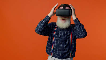 Elderly bearded gray-haired man using virtual reality futuristic technology VR app headset helmet to play simulation 3d 360 video game, drawing. Orange studio background. Senior grandfather pensionerの写真素材