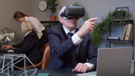 Senior male watching virtual reality 3D 360 video simulation at office. Focused business man entrepreneur using VR app headset helmet doing research. Busy freelancer working on modern tech deviceの写真素材