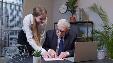 Handsome joyful senior businessman examining financial data and discussing new project with young happy woman colleague in office. Celebrating good financial results of successful teamwork strategyの写真素材