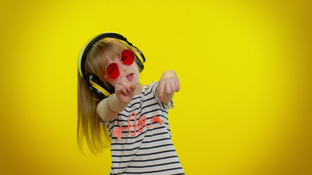 Funny playful blonde kid child 5-6 years old listening music via headphones, dancing disco fooling around having fun expressive gesticulating hands on yellow studio background. Teenager children girlの写真素材