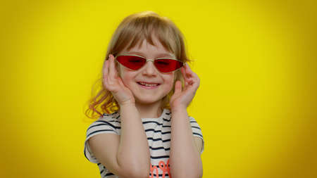 Little cute teen kid child girl in red stylish sunglasses listening music dancing disco fooling having fun expressive gesticulating hands on yellow studio background. Young children lifestyle emotionsの写真素材