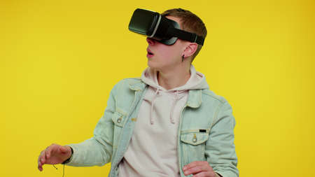 Amazed teen student boy using headset helmet app to play simulation game. Watching virtual reality 3D 360 video. Young man in VR goggles isolated on yellow background. Addiction from technologyの写真素材