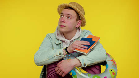 Displeased tired boring traveler tourist teen stylish boy in summer clothes on yellow background. Passenger traveling on weekends. Air journey trip delay. Hold suitcase, tickets waiting for flightの写真素材