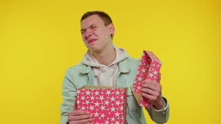 Handsome stylish teen boy unwrapping gift and expressing disappointment, dislike to bad present, guy dissatisfied with birthday surprise. Young adult man indoor isolated on yellow studio backgroundの写真素材