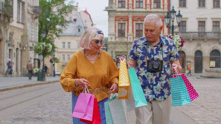 Senior old stylish tourists man woman walks with colorful bags after shopping in mall. Elderly grandmother, grandfather having good time, traveling together on city street Lviv, Ukraine. Mature familyの写真素材