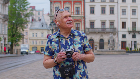 Senior man tourist with retro photo camera, smiling, listening music on earphones, dancing on summer city street center of Lviv, Ukraine. Photography, travelling vacation. Active life after retirementの写真素材