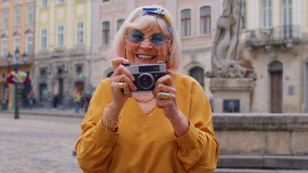 Portrait of senior woman tourist taking pictures with photo camera, smiling using retro device in summer city center of Lviv, Ukraine. Active life after retirement. Photography, travelling, vacation.の写真素材