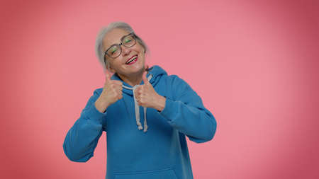 Mature old granny gray-haired grandmother in casual blue hoodie showing thumbs up and nodding in...