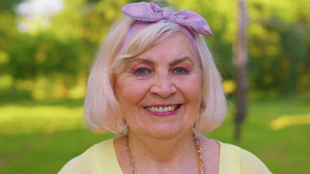 Cheerful lovely senior old gray-haired woman in casual yellow blouse posing isolated on summer park...