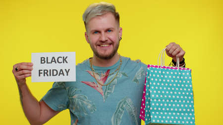 Happy tourist stylish man showing Black Friday inscription banner text, advertising discounts, smiling looking amazed with low prices, shopping on big sale day. Young guy on yellow studio backgroundの写真素材