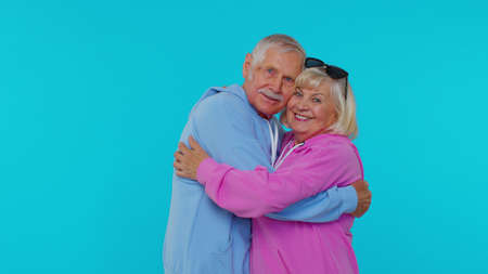 Happy cheerful senior couple man woman grandparents in casual hoodie posing isolated on blue studio background. Elderly grandfather grandmother smiling hugging enjoying successful lifestyle togetherの写真素材