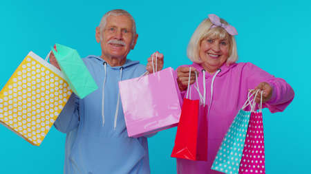 Elderly woman grandmother with man grandfather showing shopping bags, advertising discounts, smiling looking amazed with low prices, Black Friday holidays. Senior granny, grandpa on blue backgroundの写真素材