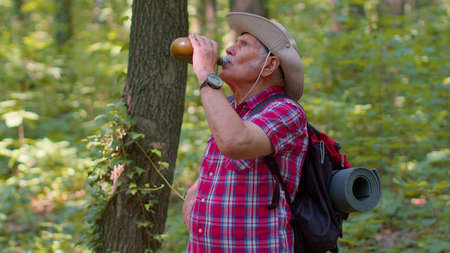 Tired thirsty old Caucasian hiker grandfather relaxing, drinking water in forest after walk. Senior...