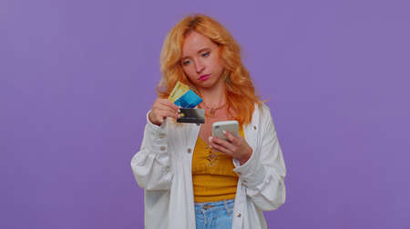 Sincere redhead lovely girl 20s years old in white shirt using credit bank cards and smartphone while transferring money purchases online shopping. Young adult woman isolated on purple wall backgroundの写真素材