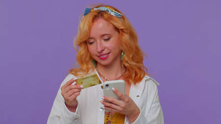 Sincere redhead lovely girl 20s years old in white shirt using credit bank card and smartphone while...