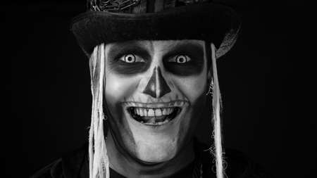 Close-up shot of creepy man in skeleton makeup. Guy opening his eyes and looking spooky at camera. Voodoo rituals. Baron Saturday. Halloween thematic party. Black background. Black and white portraitの写真素材