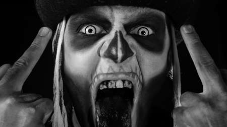 Sinister man with skull makeup making faces and showing middle finger. Bad manner gesture. Scary guy in thematic carnival costume of Halloween skeleton against dark black background. Black and whiteの写真素材