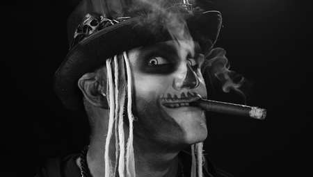 Creepy man with skeleton makeup in top-hat with feathers. Guy smoking cigar, making faces, looking at camera, smiling. Voodoo rituals. Halloween thematic party. Alone on Black backgroundの写真素材