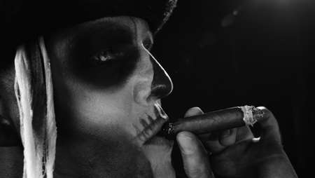 Frightening man in skeleton Halloween cosplay costume. Crazy guy in creepy skull makeup smoking cigar, making faces. Day of The Dead. Alone on black background. Black and white portraitの写真素材