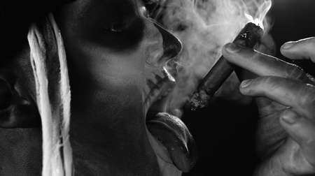 Close-up shot of frightening man in skeleton Halloween costume. Crazy guy in creepy skull makeup smoking cigar, making faces showing tongue. Day of The Dead. Alone on black background. Black and whiteの写真素材