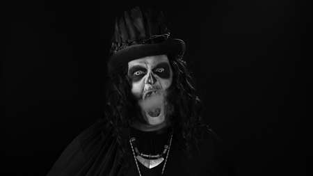 Sinister man with horrible Halloween skeleton makeup in costume with top-hat exhaling cigarette...