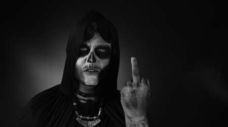 Sinister man with skull makeup making faces and showing middle finger. Bad manner gesture. Scary guy in thematic carnival costume of Halloween skeleton against dark background. Black white portraitの写真素材