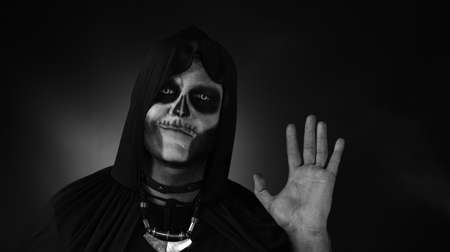 Frightening man in skeleton Halloween cosplay costume. Guy in creepy skull makeup waving hands looking at camera with smile, saying hi, hello, greeting. Day of The Dead. Black and white portraitの写真素材