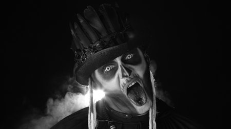 Sinister, crazy man with skeleton makeup in top-hat. Guy making angry faces, shaking head, showing tongue. Voodoo rituals. Baron Saturday. Halloween party. Black background. Black and white portraitの写真素材