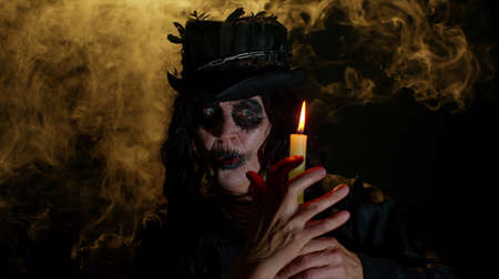 Portrait of sinister senior old mature woman in carnival costume of Halloween witch. Voodoo magic rituals with candle isolated on black studio background. Horror theme of cosplay clown, vampire, beastの写真素材