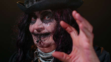 Creepy mature woman grandmother with Halloween stylish witch makeup. Old scary granny making faces, looks at camera and smiles terribly. Voodoo rituals. Thematic party. Sinister senior lady vampireの写真素材