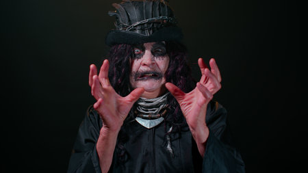 Frightening senior woman with Halloween witch makeup looking at camera, trying to scare, face expressions. Horror theme. Sinister lady on studio black background. Voodoo rituals. Fashion body artの写真素材