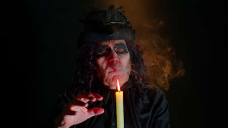 Sinister old elderly man with horrible scary Halloween witcher makeup in costume looking ominous at camera, making voodoo magic rituals with candle. Vampire grandpa isolated against black backgroundの写真素材