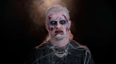 Sinister man with horrible scary Halloween zombie makeup in costume making faces, looking ominous at camera, trying to scare. Dead guy with wounded bloody scars face isolated against black backgroundの写真素材